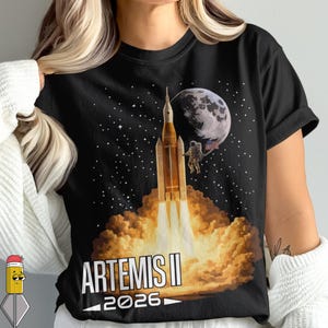 Peut inclure: T-shirt noir avec un motif spatial. Le design comprend une fusée en lancement avec des flammes, un astronaute, la lune et des étoiles. Le texte "ARTEMIS II 2026" est imprimé en bas.