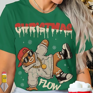 Peut inclure: T-shirt vert avec un motif sur le thème de Noël. Le design présente un bonhomme en pain d'épices portant une casquette rouge, des lunettes de soleil et une chaîne en or, dans une pose de breakdance. Le mot "CHRISTMAS" est au-dessus du bonhomme en pain d'épices, et "FLOW" est en dessous.