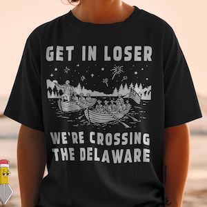Peut inclure: T-shirt noir avec un motif blanc représentant une scène historique de personnes en bateaux traversant une rivière, avec le texte "GET IN LOSER" au-dessus et "WE'RE CROSSING THE DELAWARE" en dessous.