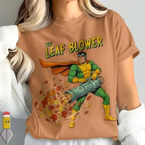 Peut inclure: T-shirt de couleur beige avec une illustration de style bande dessinée d'un super-héros utilisant un souffleur de feuilles pour expulser des feuilles d'automne. Le texte "THE LEAF BLOWER" est au-dessus du super-héros. Le super-héros porte un costume vert et jaune avec une cape orange.