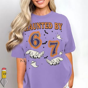 Peut inclure: T-shirt lilas avec l'inscription "HAUNTED BY" en orange au-dessus des chiffres "6" et "7". Les chiffres sont soulignés en bleu marine. Le t-shirt présente des chauves-souris, des fantômes et des mains.