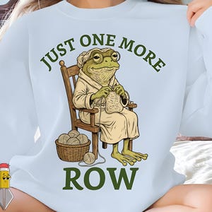 Peut inclure: Sweat-shirt bleu clair avec une grenouille de dessin animé portant des lunettes et une robe, tricotant dans un fauteuil à bascule en bois. Le texte "JUST ONE MORE ROW" est arqué au-dessus et en dessous de la grenouille.
