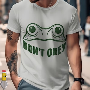 May include: A pale grey t-shirt with a green frog face design and the words "DON'T OBEY" in a bold font. The frog's eyes are large and round. The shirt is a short-sleeved crew neck.