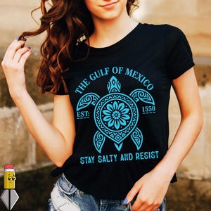 Peut inclure: T-shirt noir avec un motif de tortue turquoise. Le motif comprend le texte "THE GULF OF MEXICO", "EST: 1550" et "STAY SALTY AND RESIST". La tortue a un motif floral au centre.