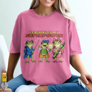 Peut inclure: T-shirt rose avec l'inscription "TEACHING IS MY SUPERPOWER" et un dessin de trois grenouilles de dessin animé habillées en professeurs. Elles portent un blazer bleu, un gilet vert et une jupe rose.
