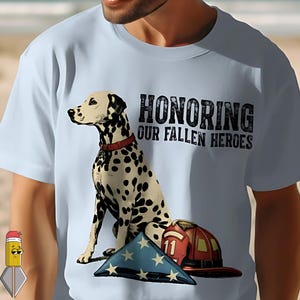 Peut inclure: T-shirt bleu clair avec un dalmatien, un drapeau américain plié et un casque de pompier. Le texte "HONORING OUR FALLEN HEROES" est imprimé au-dessus du chien. Le chien porte un collier rouge.