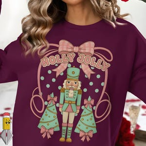 Peut inclure: Un sweat-shirt bordeaux avec un motif représentant un casse-noisette, des sapins de Noël et le texte "HOLLY JOLLY". Le motif comprend des nœuds roses et des accents turquoise, créant un thème de vacances festif.