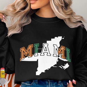 Peut inclure: Sweat-shirt noir avec un contour blanc d'une carte de ville et le mot "MIAMI" en lettres orange et vertes. Le sweat-shirt est de couleur noire unie.