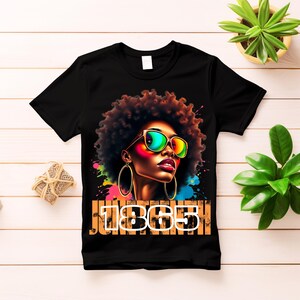 Può includere: Maglietta nera con un'illustrazione colorata di una donna con occhiali da sole e grandi orecchini a cerchio. Il testo "Juneteenth 1865" è stampato sulla maglietta.