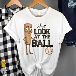 Op de afbeelding: Wit t-shirt met een cartoon houten figuur en de tekst "Just LOOK AT THE BALL" en een honkbal. De figuur wijst naar de bal. Het t-shirt hangt aan een houten hanger.
