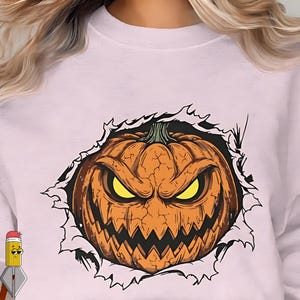 May include: A light pink sweatshirt featuring a Halloween-themed graphic. The graphic depicts a carved orange pumpkin with a menacing face, bursting through a torn paper effect. The pumpkin has yellow eyes and a jagged mouth.