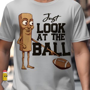 Op de afbeelding: Lichtgrijs t-shirt met een cartoon houten figuur dat naar een American football wijst. De tekst "Just LOOK AT THE BALL" is in het zwart gedrukt. De cartoonfiguur heeft een lachend gezicht.