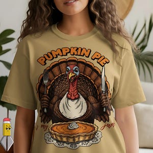 Peut inclure: T-shirt de couleur beige avec un dessin de dinde de dessin animé tenant une fourchette et un couteau, avec une tarte à la citrouille sur une nappe dentelle. Le texte "PUMPKIN PIE" est au-dessus de la dinde, et "my jam" est écrit en dessous.