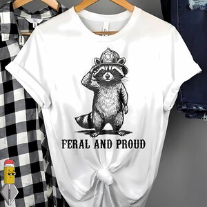 Peut inclure: T-shirt blanc avec une illustration en noir et blanc d'un raton laveur portant un casque de pompier et saluant. Le texte "FERAL AND PROUD" est imprimé en dessous.