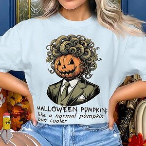 May include: Light blue t-shirt featuring a Halloween-themed graphic. The design depicts a pumpkin head with curly hair, wearing a suit and tie. Text on the shirt reads: "HALLOWEEN PUMPKIN; like a normal pumpkin but cooler."