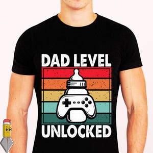 Peut inclure: T-shirt noir avec un motif graphique représentant un biberon en forme de manette de jeu vidéo. Le texte "DAD LEVEL UNLOCKED" est au-dessus et en dessous du motif de la manette, sur un fond rayé rouge, orange, jaune, turquoise et vert.