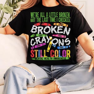 May include: Black t-shirt with the text "We're all a little broken, but the last time I checked, broken crayons still color." and "Mental Health Awareness" below. The text is surrounded by colorful broken crayon images.