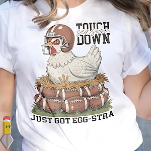 May include: White t-shirt featuring a cartoon chicken wearing a football helmet, sitting on a nest of footballs. Text reads "Touchdown" and "Just Got Egg-stra". The design is a humorous take on football season.