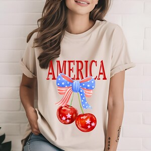 Peut inclure: Un t-shirt beige avec un graphique "AMERICA" rouge et un nœud rouge, blanc et bleu avec des étoiles au-dessus de deux cerises rouges avec des étoiles blanches.
