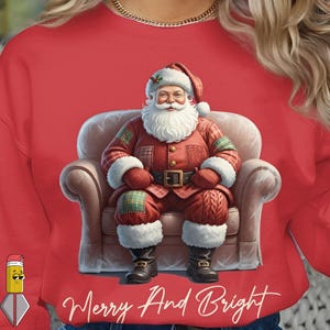 Peut inclure: Sweat-shirt rouge avec une illustration numérique du Père Noël assis dans un fauteuil. Il porte un costume rouge avec des bordures blanches et un chapeau assorti. Le texte "Merry And Bright" est écrit en cursive en bas.