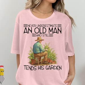 Op de afbeelding: Lichtroze t-shirt met de tekst "NEVER UNDERESTIMATE AN OLD MAN WHO STILL TENDS HIS GARDEN." De afbeelding toont een illustratie van een persoon die aan het tuinieren is, met een hoed en blauw overhemd, omringd door planten en potten.