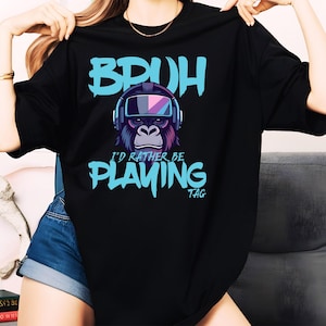 Peut inclure: T-shirt noir avec un graphique bleu d'un gorille portant des lunettes VR et le texte "Bruh I'd rather be playing TAG".