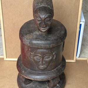 May include: A dark brown, carved wooden sculpture featuring a stylized human figure atop a cylindrical base. The sculpture has a weathered appearance, with visible details including facial features and a textured head covering. The base has a carved face.