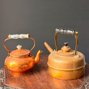 May include: A copper kettle with a white ceramic handle featuring blue floral patterns and a matching lid knob, positioned next to a brass kettle with a black handle and a cylindrical body, both displaying a metallic finish and patina.