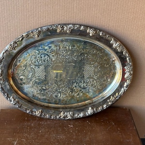 May include: Oval silver-plated serving tray with ornate floral and scrollwork detailing. The rim features a grape cluster design. The tray is approximately 18 inches long and 12 inches wide, with a slightly tarnished appearance.