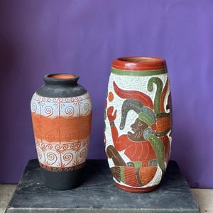 May include: Two ceramic vases with intricate designs. The vase on the left has a black base, orange and white bands with spiral patterns. The taller vase features a detailed relief of a figure in red, green, and brown against a white background.