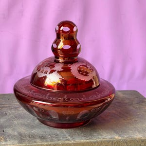 May include: A deep red glass candy dish with a matching lid, featuring intricate etched floral and architectural patterns. The lid is topped with a tiered, circular finial with cut-out details, and the base has a wide, rounded bowl shape.