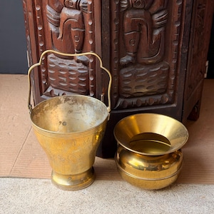May include: A brass bucket with a swing handle and etched floral patterns alongside a smaller, smooth brass bowl with a wide, flared rim. Both items are metallic gold in color and feature a polished finish, showing signs of natural aging and patina.
