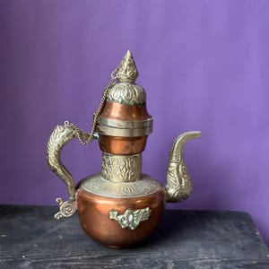 May include: An antique copper and brass teapot with ornate detailing. The teapot features a curved spout, a decorative handle, and a hinged lid with a pointed finial. A small chain connects the lid to the handle. The teapot is displayed on a dark wooden surface against a purple background.