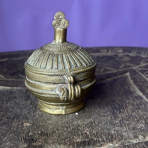 May include: A small, antique brass container with a lid. The container has a round body with detailed etched patterns and a decorative handle. The lid is topped with a swirl design. The container is displayed on a wooden surface.