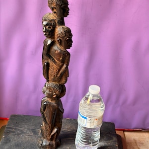 May include: A dark brown wooden sculpture featuring a vertical stack of four carved human figures, each with detailed facial features and textured hair, standing next to a clear plastic water bottle for a sense of scale.