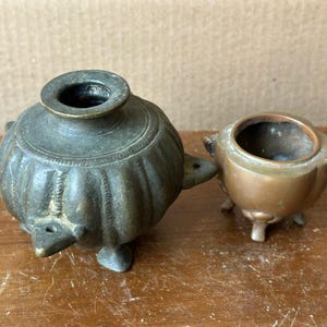May include: Two antique metal vessels. One is a dark green, bulbous pot with a small opening and three feet. The other is a smaller, bronze-colored pot with three feet and a wide opening. Both have a weathered appearance.