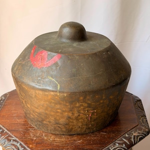 May include: A dark bronze-toned metal gong with a central raised knob. The surface features a weathered, textured patina with visible signs of wear, including a small red painted mark and a faint yellow streak on the upper side of the bowl.