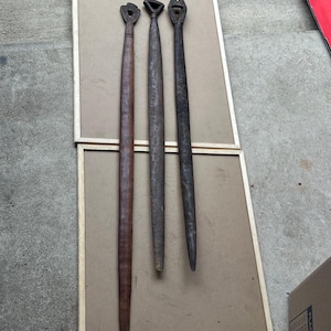 May include: Three antique wooden tools with carved handles and long, tapered shafts. The tools are displayed on a light brown surface, each with a unique handle design. The wood tones range from light brown to dark brown.