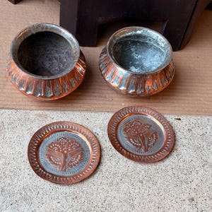 May include: Two round metal pots with fluted sides and a metallic copper and silver finish, accompanied by two matching circular metal lids or coasters featuring an embossed floral pattern in the center and a decorative border design.