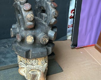 May include: A carved wooden sculpture featuring a stylized face and a geometric upper section. The sculpture is primarily dark brown with accents of gold and red. The face has detailed carvings and a light brown color.