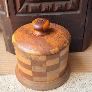 May include: A cylindrical wooden container with a lid, featuring a checkerboard pattern of alternating light and dark wood squares. The lid has a rounded knob handle and concentric circular carvings. The base is a solid, circular wooden disc.