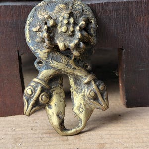 May include: A metallic gold-toned decorative object featuring two intertwined serpent-like heads at the bottom and a textured, circular floral-inspired motif at the top. The piece has a weathered, aged finish with dark recessed areas and a worn, uneven surface.