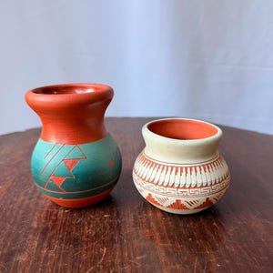 May include: Two small clay pots. The left pot is terracotta-colored with a teal band featuring etched geometric triangles. The right pot is cream-colored with a terracotta-colored rim and base, decorated with intricate etched geometric patterns.
