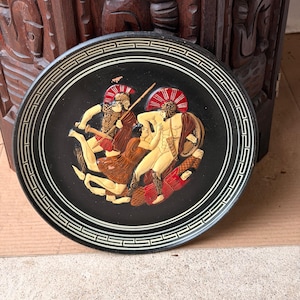 May include: A circular black decorative plate featuring a raised relief scene of two ancient warriors in combat. The figures are painted in cream, red, and brown tones. The rim is decorated with a white Greek key pattern on a black background.