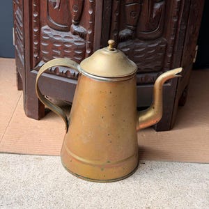 May include: A vintage brass coffee pot with a tapered cylindrical body, a curved handle, a long narrow spout, and a removable lid with a small knob. The metal surface shows signs of age, including dark patina, surface spots, and discoloration.