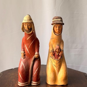 May include: Two hand-carved wooden figurines. The left figure wears a red robe, a tan head covering, and holds a staff. The right figure wears a yellow robe, a brown hat, and carries a small child in a pouch on the front of the garment.