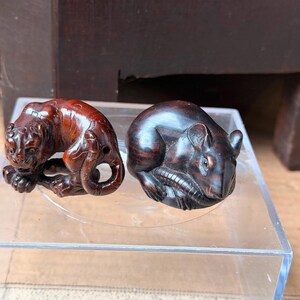 May include: Two small, hand-carved wooden figurines. One is a reddish-brown tiger in a crouching position with visible facial features and a curled tail. The other is a dark brown, smooth-surfaced mouse curled into a rounded, compact shape.