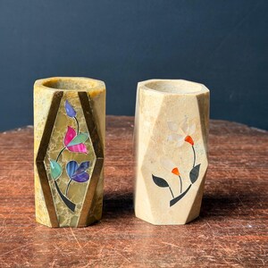 May include: Two cylindrical stone vessels with faceted sides. The left vessel is mottled yellow-green stone with a brass-framed inlay featuring colorful floral petals. The right vessel is light beige stone with a white and orange floral inlay.
