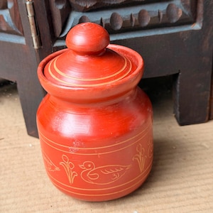 May include: A round, bright red lidded jar featuring a smooth finish and a knobbed lid. The exterior is decorated with thin, etched golden-yellow line drawings of ducks and floral motifs, framed by horizontal bands encircling the body and lid.