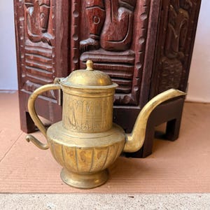 May include: A brass teapot with a rounded body, a long curved spout, and a lid featuring an engraved pattern. The teapot has a wide base, a handle on the side, and a smaller decorative handle near the bottom. The surface shows a metallic finish.